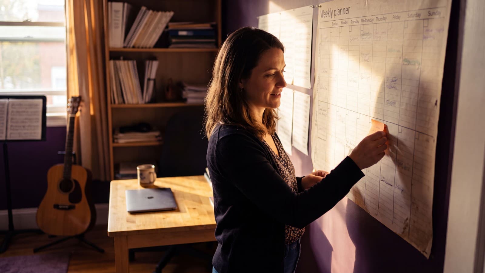Music teacher places a sticky note into a large weekly wall planner, guitar and sheet music shelf in warm afternoon light