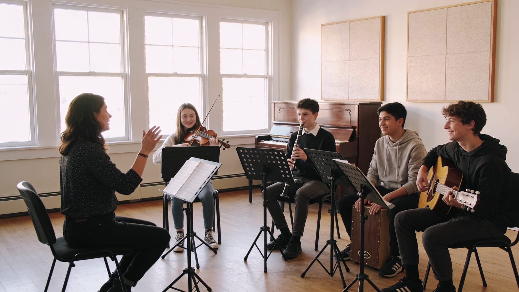 Ensemble rehearsal at a music school: a teacher cues four teenagers on violin, clarinet, cajón and guitar